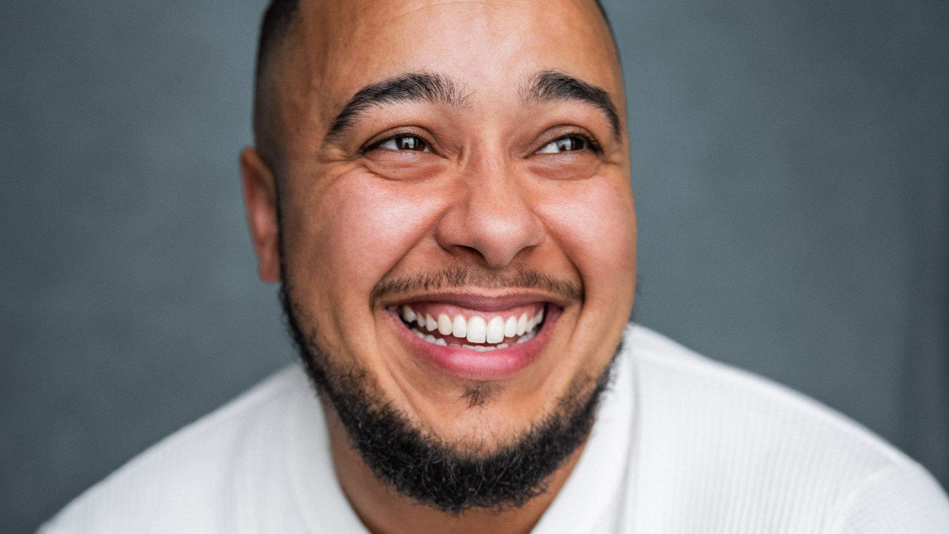 Mika is a 34 year old black mixed-race person, a trans man with short hair and a neatly trimmed beard, he smiles brightly looking off camera, showing his teeth. He wears a textured white shirt and sits against a plain grey backdrop.