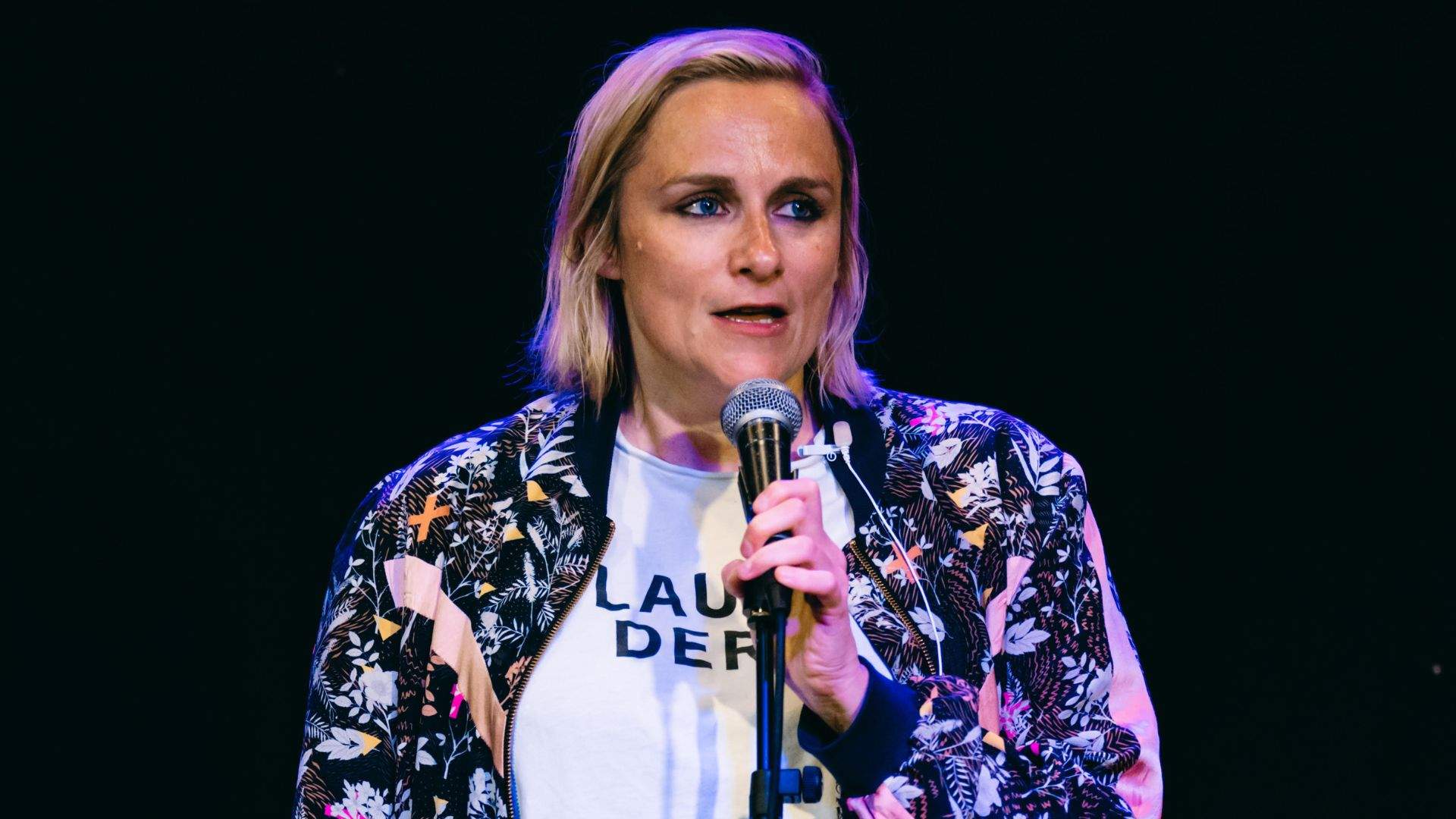 Jenny is a white woman with blonde hair and blue eyes. She is wearing a floral bomber jacket with pink stripes on the arms. She is speaking into a mic on a mic stand.