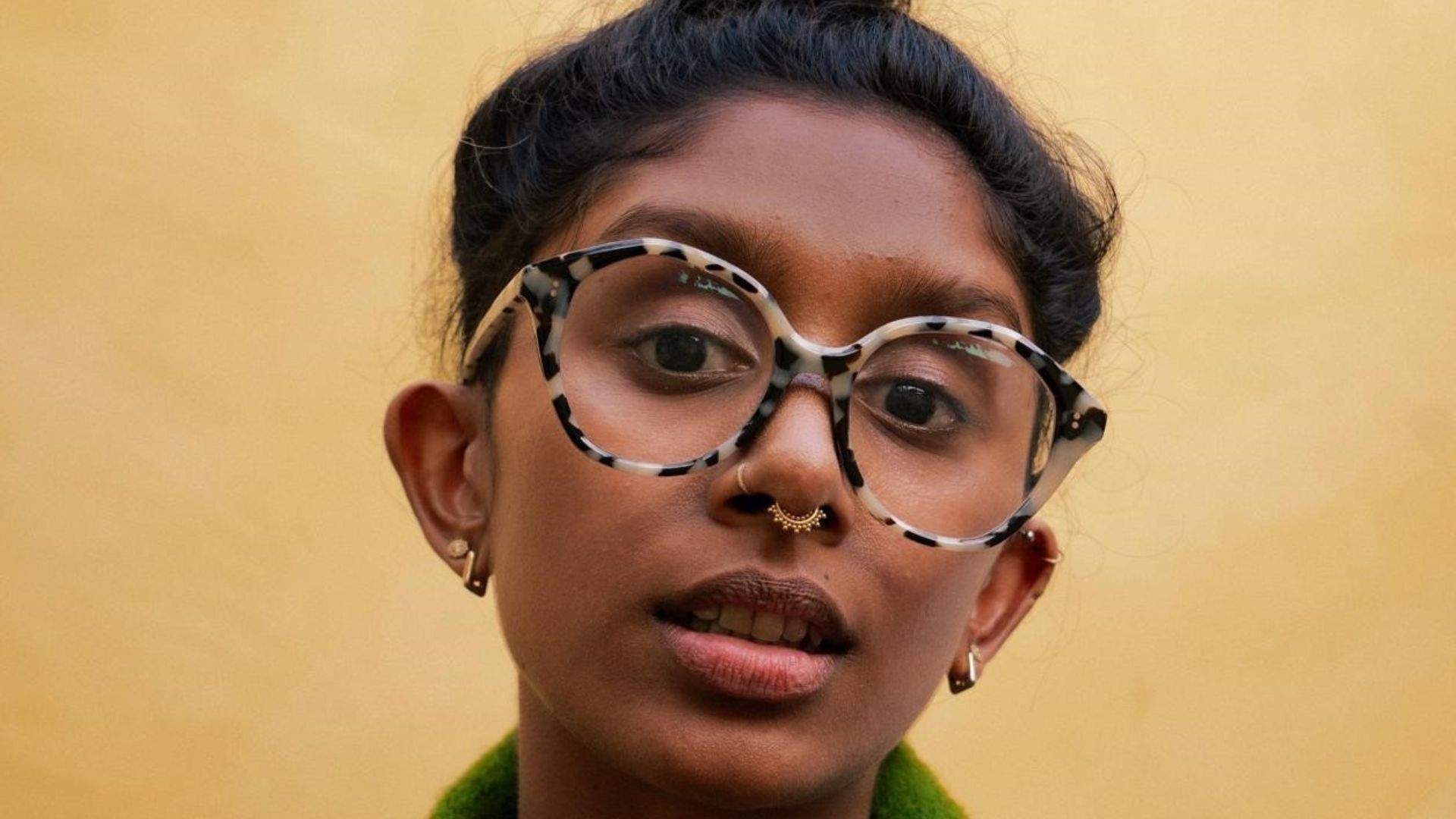 Headshot of Gayathiri, a brown-skinned Tamil person gazing directly into the lens. They’re wearing a moss green cardigan and big turtle shell glasses.