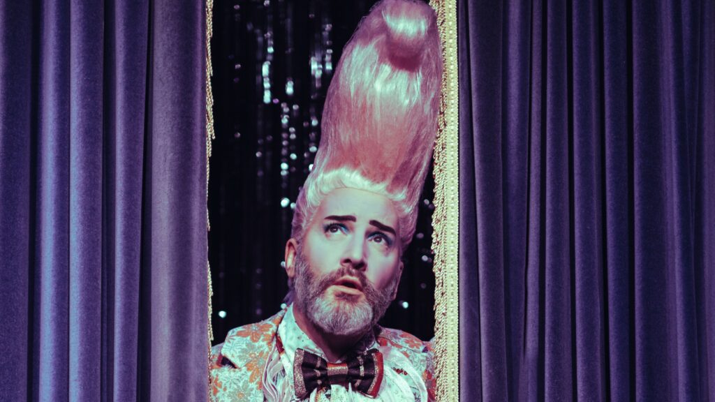 Boogaloo Stu, a white middle aged person with a big pink quiff, beard and bowtie.